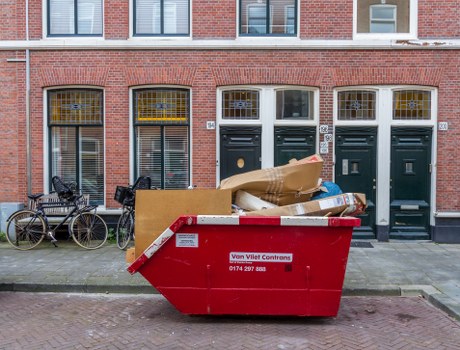 A large red commercial skip bin filled with assorted waste materials, positioned on the pavement in front of a row of brick terraced houses. The waste includes cardboard boxes, plastic bags, and miscellaneous packaging, with some items partially overflowing the bin. The skip features a white and red label indicating waste management services, and is situated directly outside the entrance doors of the residential units, which have dark green doors with large windows above. To the left of the bin, there are two bicycles parked close to the wall, one with a black frame and the other with a silver frame, leaning against the building. The background shows a typical urban street scene with brickwork, windows, and a paved walkway, suggesting an environment suitable for commercial waste removal in Brixton.