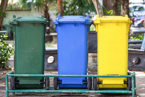 A row of three large wheelie bins positioned on a metal trolley in an outdoor urban environment, likely a pavement or residential street, with three bins in green, blue, and yellow. The bins are made of durable plastic with textured surfaces, and each has a closed hinged lid on top. The green bin, located on the left, has a slightly weathered finish, while the middle blue bin and the right yellow bin appear newer and have smooth surfaces. The backdrop includes blurred trees, park benches, and parked cars, suggesting a residential or town centre setting, possibly near Brixton. The lighting is natural daylight, illuminating the different colours and textures of the bins clearly. This scene depicts typical waste collection infrastructure, relevant to commercial waste removal services like those offered by Commercial Waste Removal Brixton, which handles various rubbish disposal needs for local properties and businesses in the area under postal code SW2.