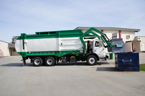 A large commercial waste collection truck with a white body and green accents is parked on a paved surface in front of a modern residential building with white walls and red window trims. The truck features a hydraulic arm mechanism extending towards a blue recycling bin positioned on the right side of the image, which is partially open, revealing black waste material inside. In the background, there are additional buildings and a clear, slightly overcast sky. The setting appears to be a residential or mixed-use area, possibly in Brixton, with the truck ready for rubbish removal services provided by Commercial Waste Removal Brixton. The surface is flat and free of debris, and the scene emphasizes the professional handling of waste collection within an urban environment, suitable for illustrating a rubbish removal service.