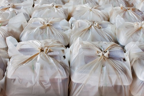 A group of large white plastic rubbish bags, tightly knotted at the top, are arranged closely together on a paved surface, likely in an outdoor urban environment such as a driveway or street in Brixton. The bags are filled with waste materials, giving them a rounded, bulky appearance with visible creases and slight transparency revealing the contents inside. The plastic finish of the bags reflects some natural light, highlighting their soft, crinkled texture. The background shows additional bags stacked or placed on the ground, creating a cluttered scene typical for rubbish removal operations in busy residential or commercial areas. This image emphasizes waste collection and disposal activities, which Commercial Waste Removal Brixton provides for local businesses and residents, fitting the context of professional rubbish management services in the Brixton area, inferred from the postcode and local town environment. The focus remains solely on the bags and their immediate surroundings, indicating a routine rubbish collection task in the borough.