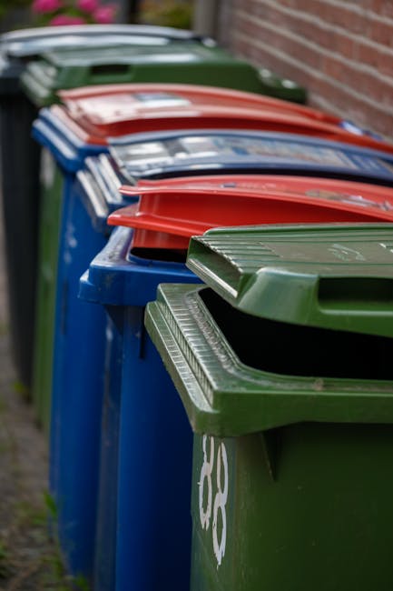 A row of large wheeled rubbish bins positioned along a pavement next to a brick wall. The bins are made of sturdy plastic with textured lids and are in various colours, including green, blue, red, and