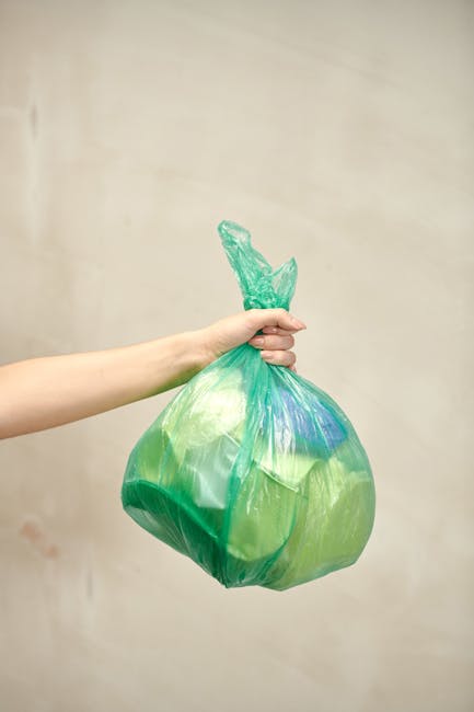 A row of large wheeled rubbish bins positioned along a pavement next to a brick wall. The bins are made of sturdy plastic with textured lids and are in various colours, including green, blue, red, and